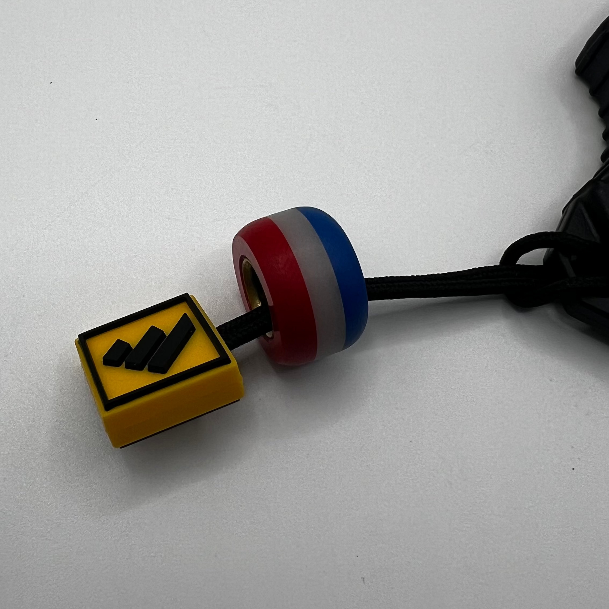 A close-up of a small yellow square charm with a black zigzag symbol, attached to a black cord and a red, white, and blue cylindrical bead, on a plain light background.