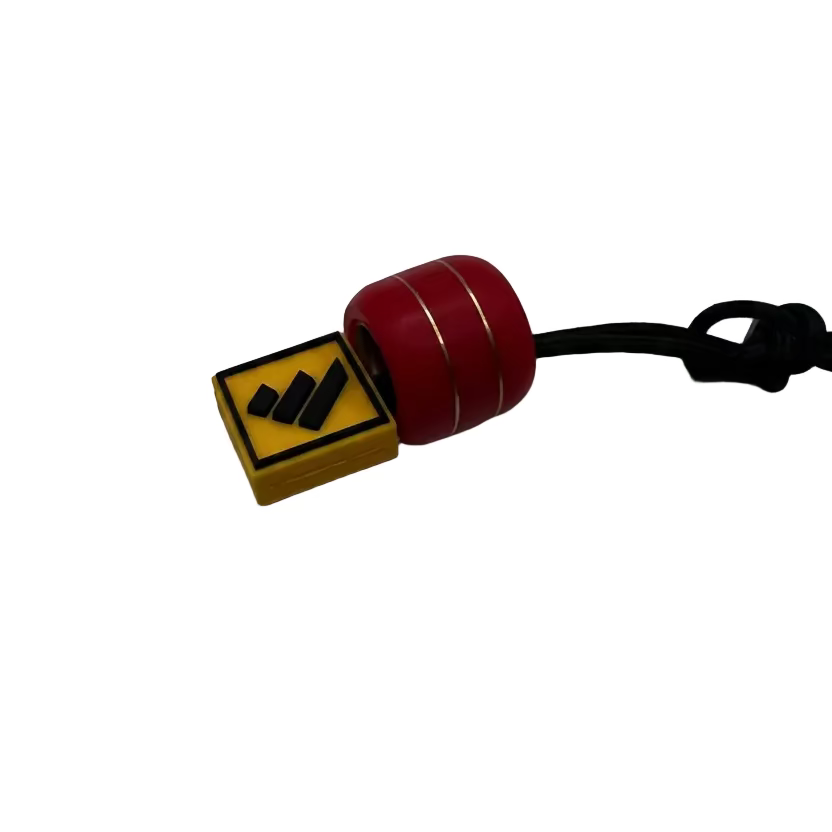 A small red cylindrical bead with thin gold stripes and a yellow square charm featuring a black graphic, attached to a black cord on a plain light gray background.