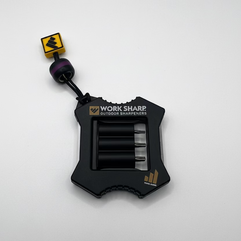 Work Sharp Outdoor Sharpeners pocket tool with a black plastic body, three sharpening wheels visible, a small yellow square key, and a purple bead on a cord, shown on a light gray background.