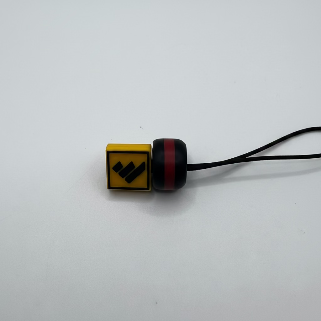 A small black and red cylindrical accessory with a yellow square end featuring a black stylized logo, attached to a black cord on a plain light background.
